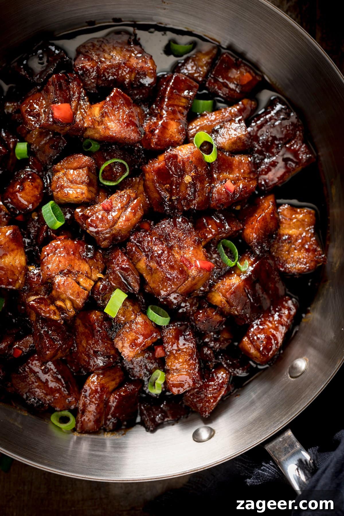 Close up overhead image of sticky Chinese pork belly, topped with chillies and spring onions in a silver frying pan, ready to serve.
