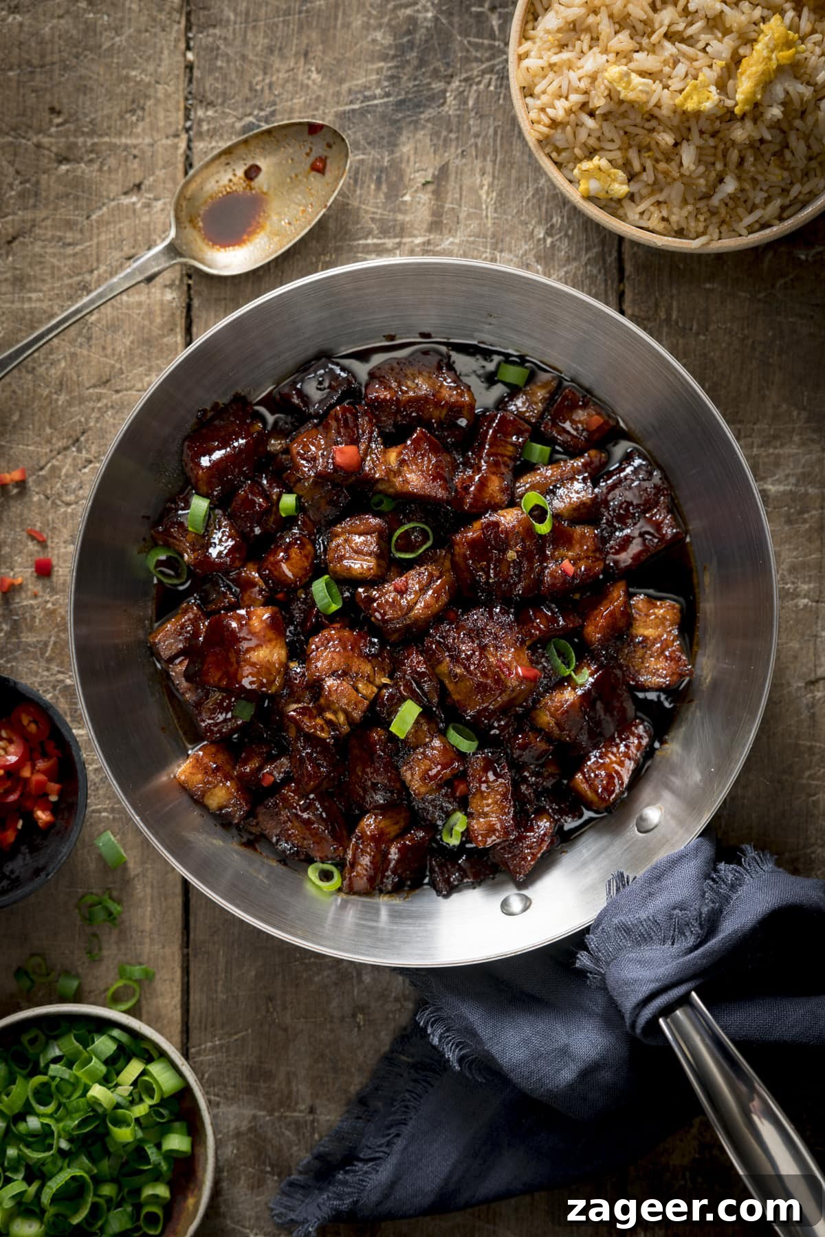 Overhead image of sticky Chinese pork belly in a silver frying pan, garnished with chillies and spring onions. The pan is on a wooden table and there are bowls of spring onions, chillies and fried rice around the pan.