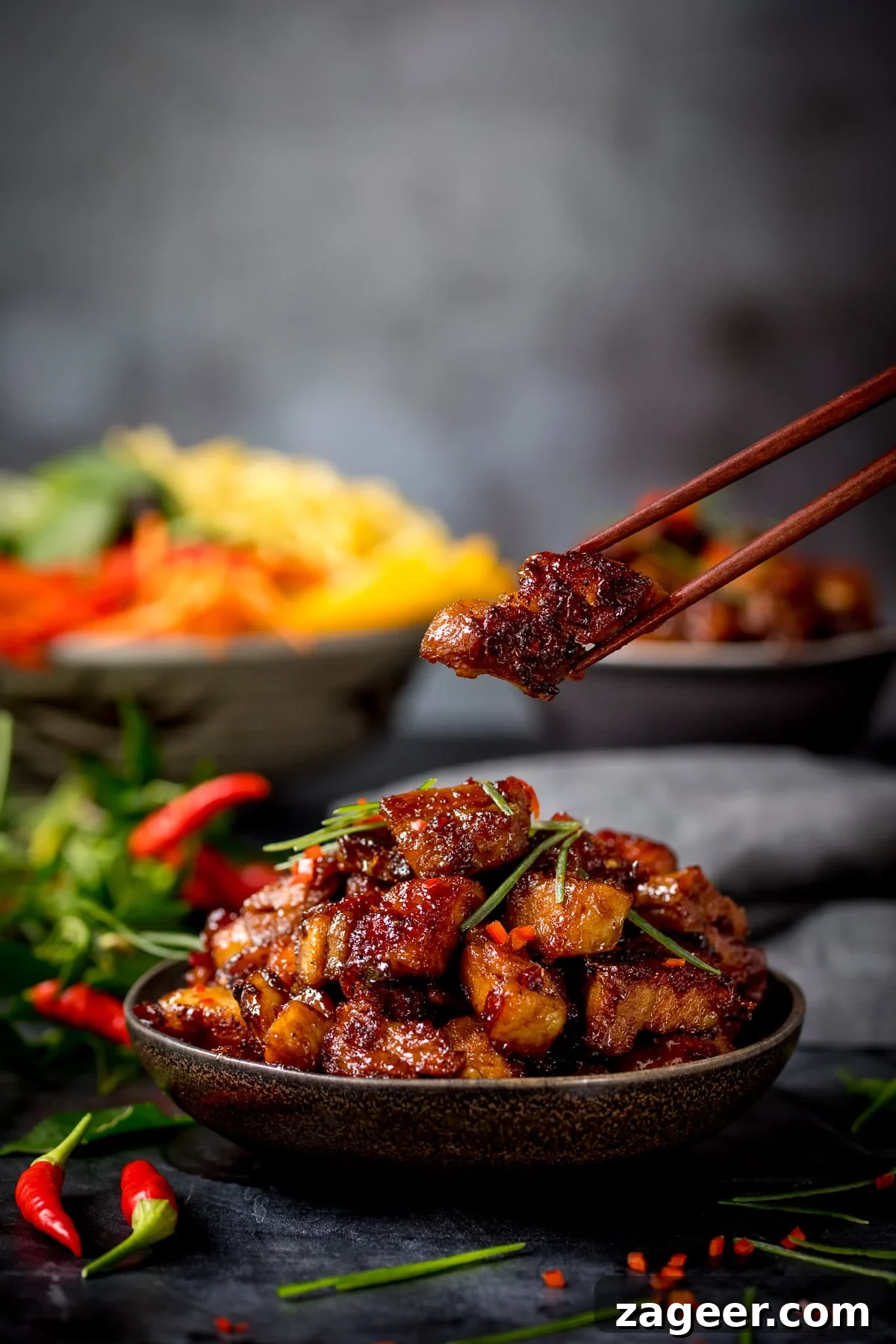 A bowl of Chinese sticky pork belly pieces against a dark background. There is a piece being lifted from the bowl with a pair of wooden chopsticks. There are ingredients scattered around and a bowl of colourful vegetables in the background.