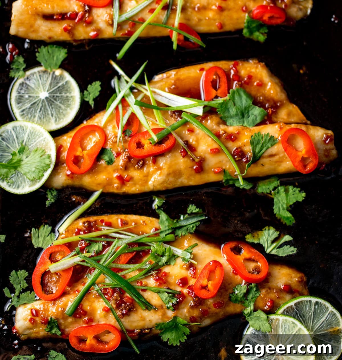 12-Minute Sweet and Sticky Asian Sea Bass 2 Baked sea bass fillets topped with sliced red chilli, spring onions, and coriander, garnished with lime slices on a dark baking tray.