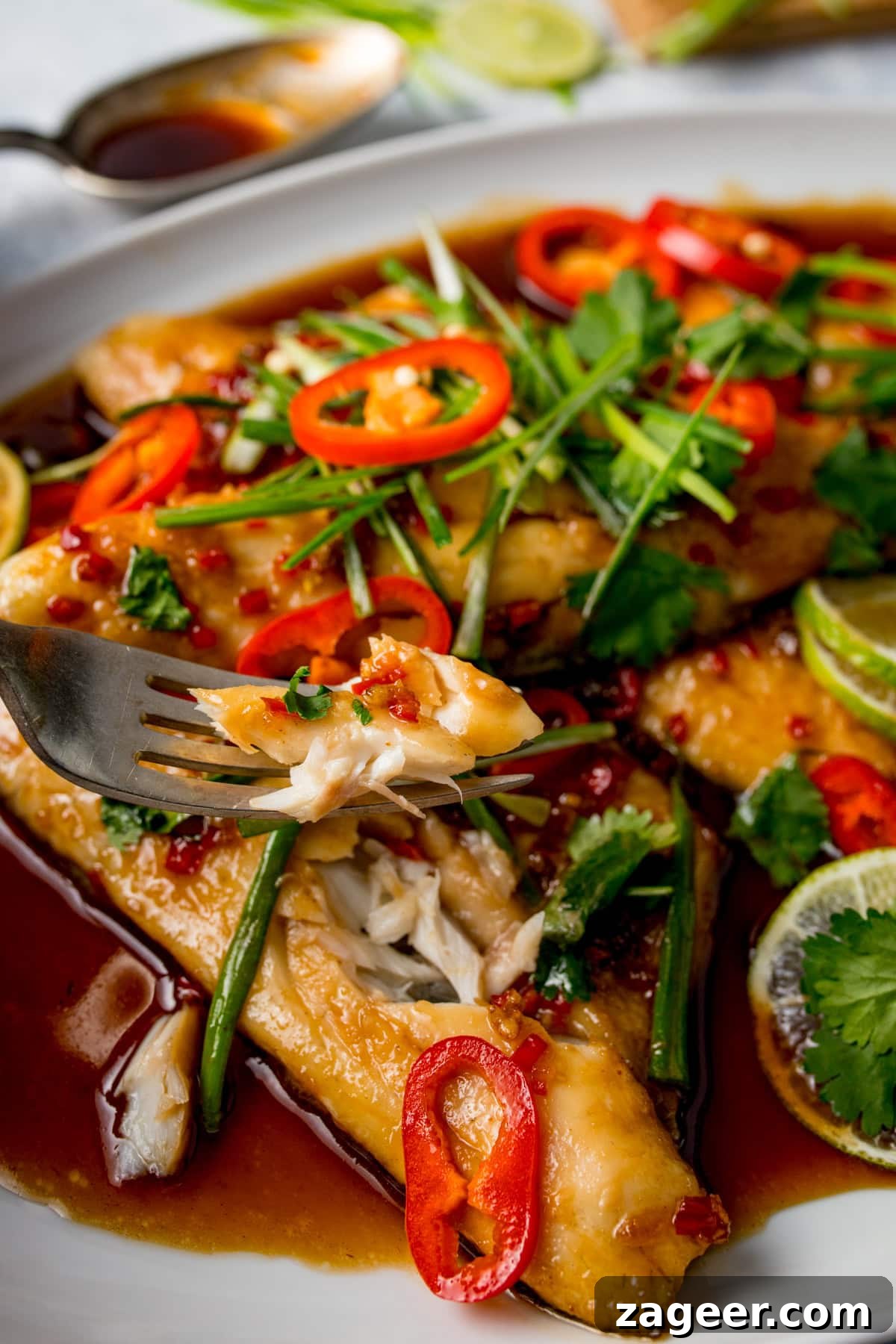 12-Minute Sweet and Sticky Asian Sea Bass 8 A close-up of Chinese Sea Bass fillets, oven baked and topped with sliced red chilli, herbs, and spring onions in a soy-based sauce, with a fork lifting a flaky piece.