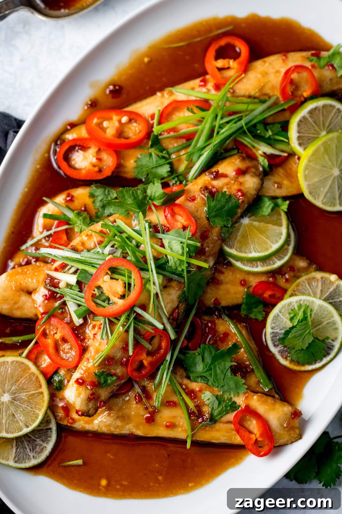 12-Minute Sweet and Sticky Asian Sea Bass 6 Oven-baked sea bass fillets topped with sliced red chilli, lime, coriander, and spring onions, served with a sticky glaze on a white platter.