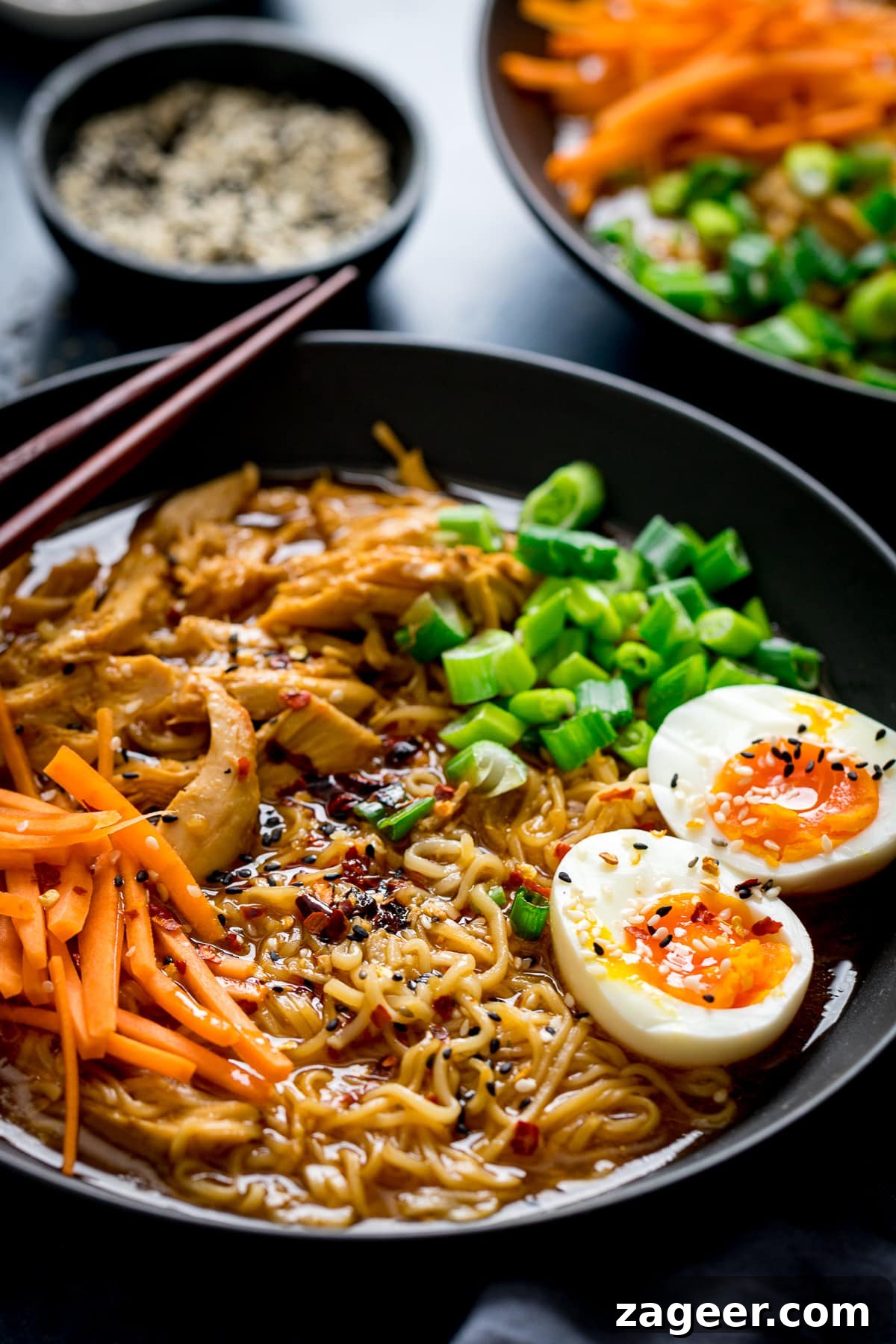 Effortless Chicken Ramen in 20 Minutes 5 A close-up of a bowl of chicken ramen, with another bowl and a small dish of sesame seeds in the soft-focus background, highlighting the egg topping.