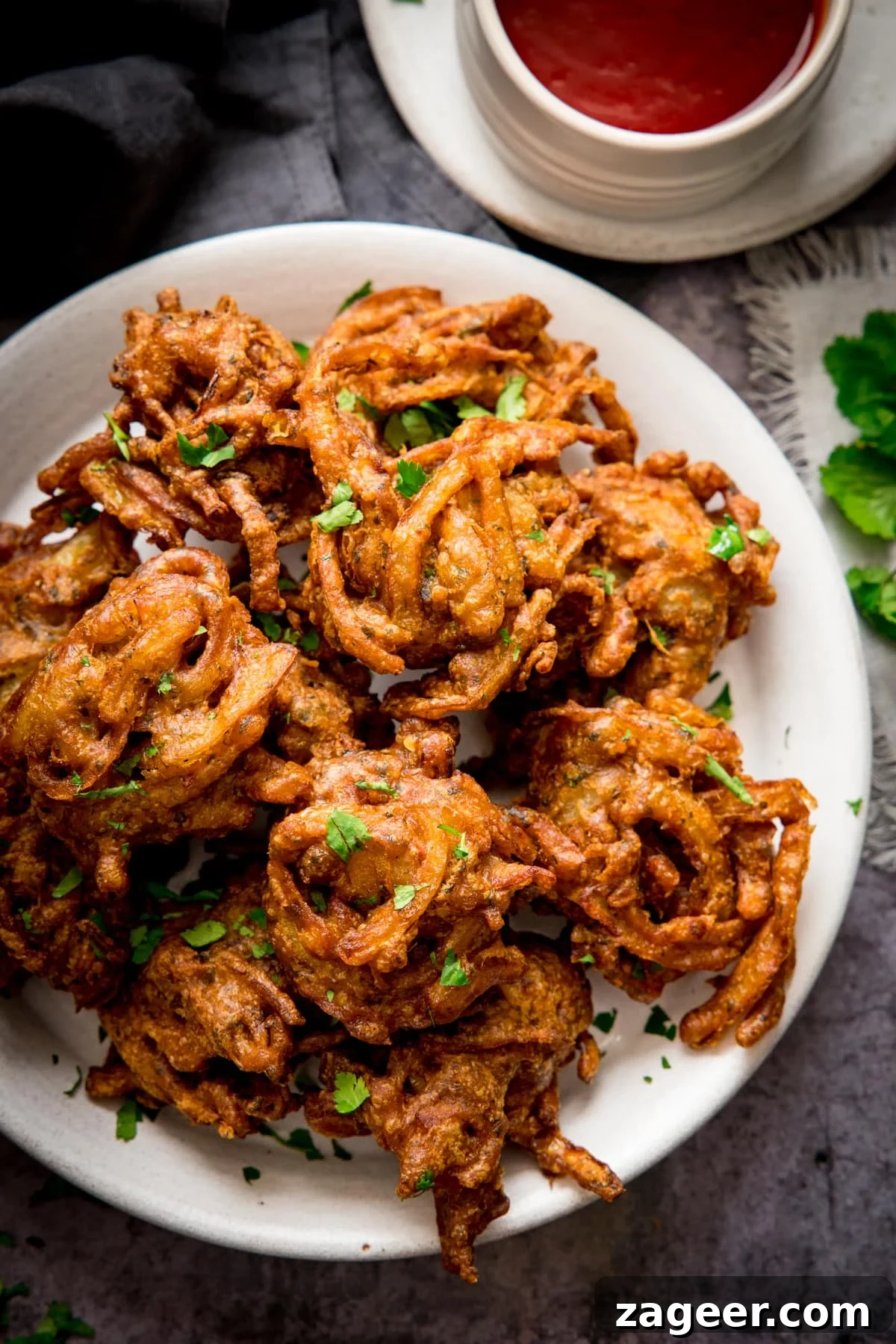 Crispy Golden Onion Bhaji 5 Crispy Onion Bhajis on a white plate with a small bowl of sweet chilli sauce in the background.