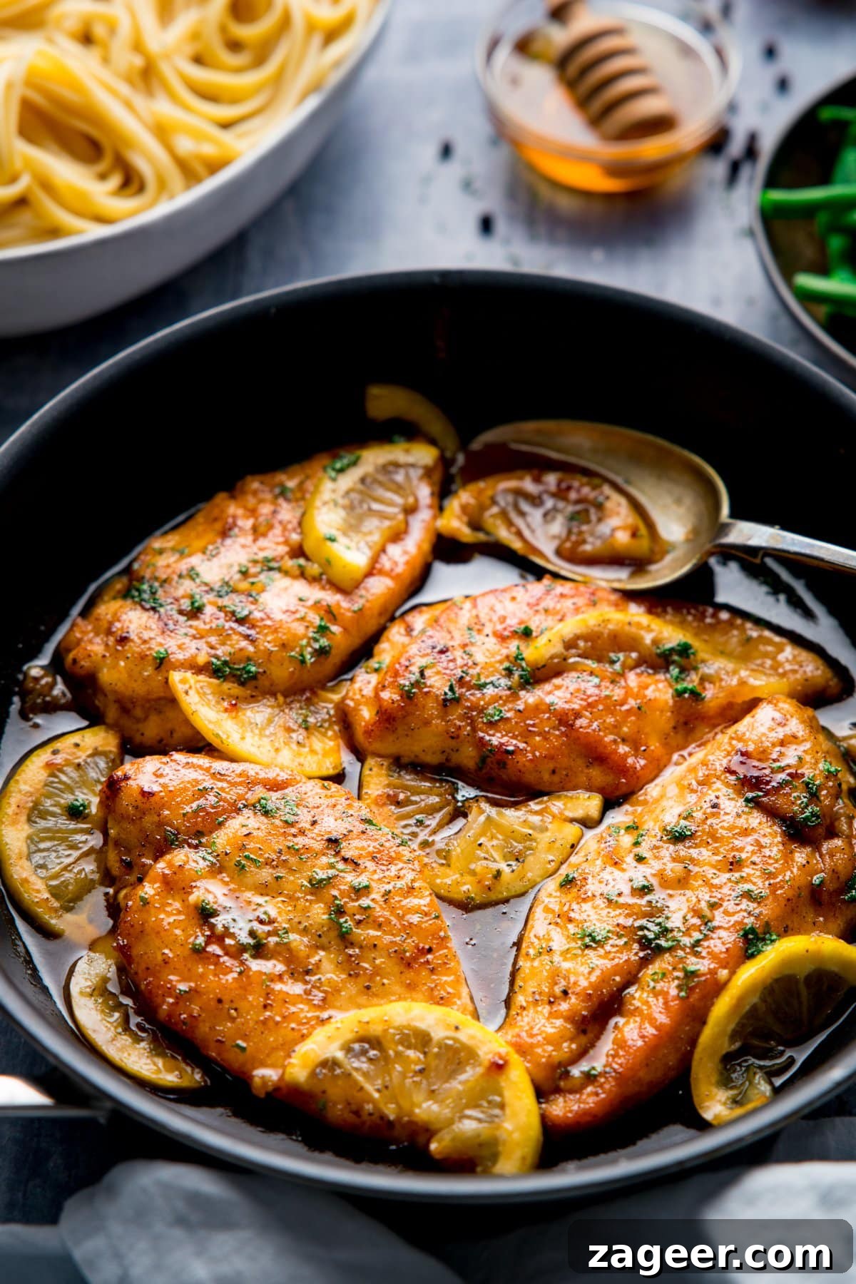 Zesty Honey Glazed Chicken 2 A dark frying pan brimming with golden honey lemon chicken pieces, garnished with fresh parsley and lemon slices, resting on a rustic blue background. A bowl of pasta and a small dish of honey are subtly visible, hinting at the dish's delightful accompaniments and flavors.