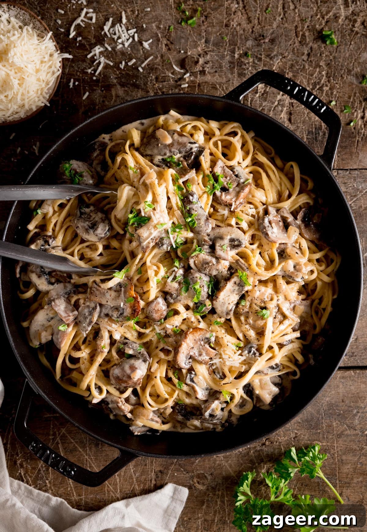 Velvety Mushroom Pasta 4 A large frying pan filled with creamy mushroom pasta, adorned with fresh parsley and surrounded by scattered grated Parmesan on a wooden surface.