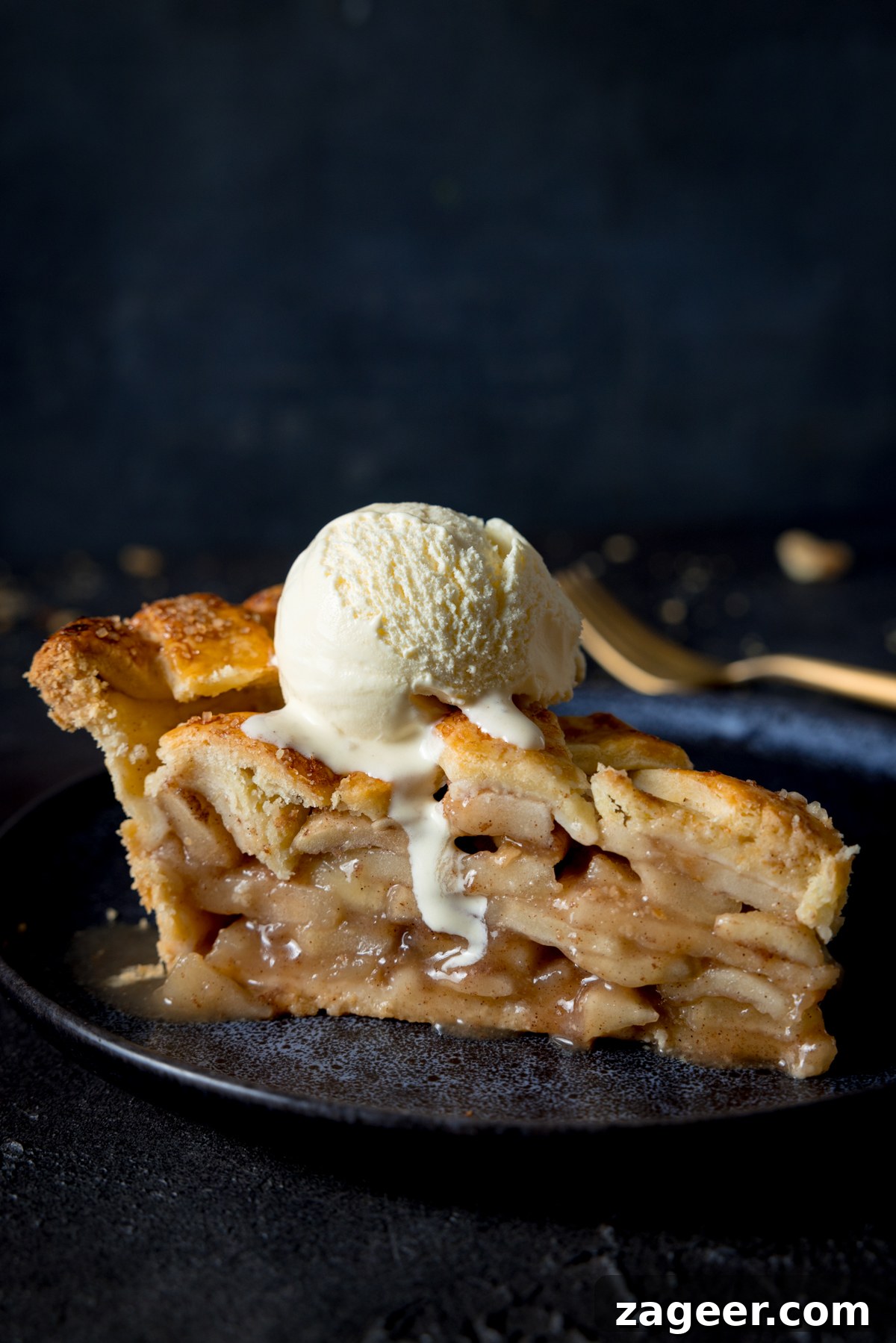 Sumptuous Deep Dish Apple Pie 2 A side shot of a slice of deep dish apple pie with a scoop of vanilla ice cream on top, on a blue plate with a napkin and fork in the background on a grey table. This image highlights the deep layers of apple filling and the golden crust.