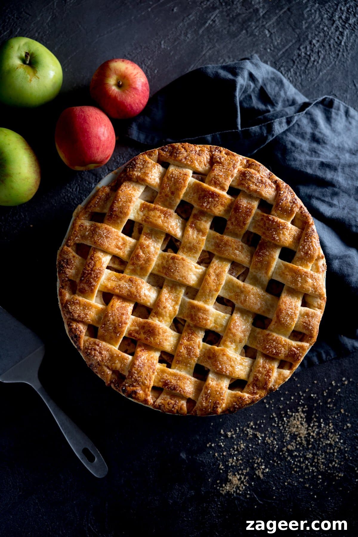 Sumptuous Deep Dish Apple Pie 4 A photo looking down on a perfectly round golden brown lattice topped deep dish apple pie, with fresh apples in the background, a blue tea towel and a spoon next to the dish. The intricate lattice crust is visible.