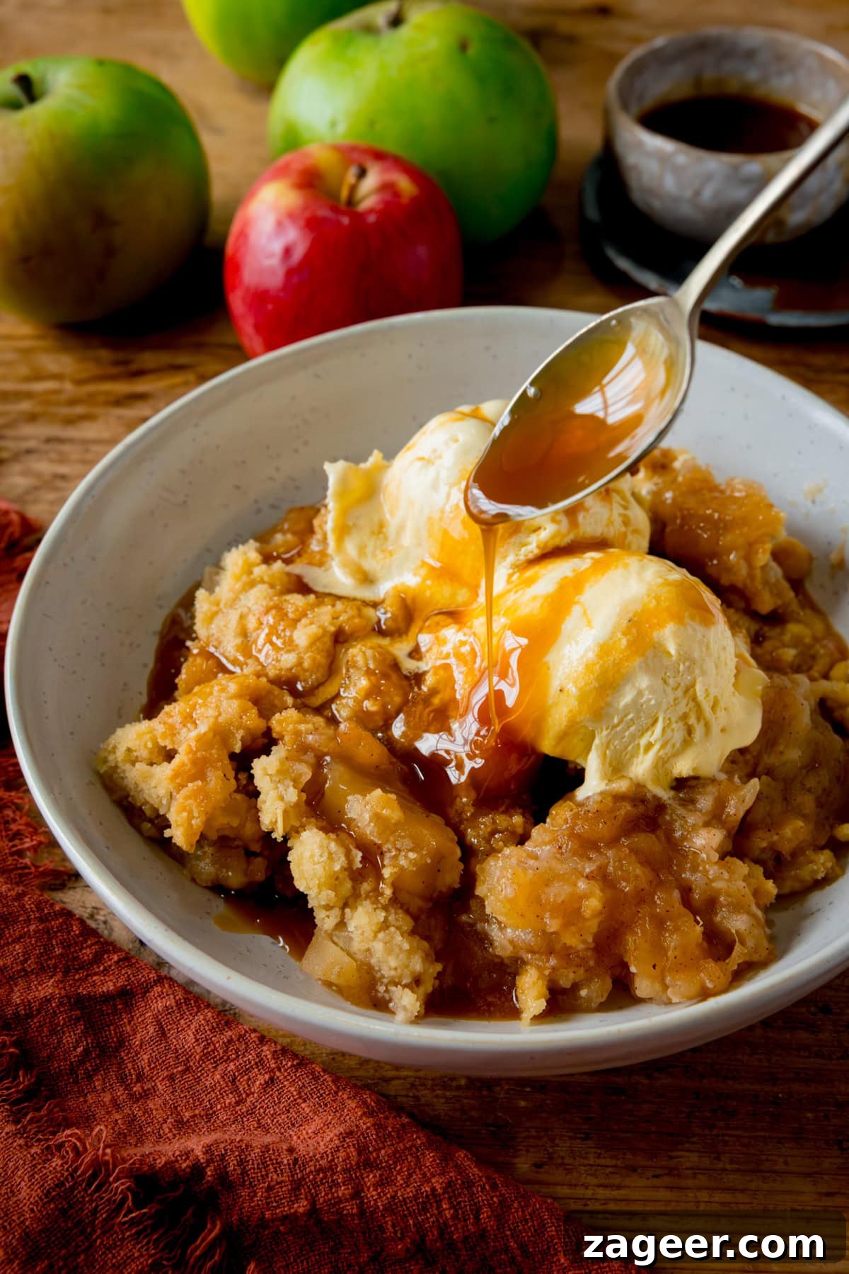 Irresistible Apple Crumble Perfection 2 A tall image of a white bowl filled with freshly baked apple crumble, generously topped with two scoops of creamy vanilla ice cream. A stream of rich caramel sauce is being drizzled over the top with a spoon, creating an irresistible finish. The bowl rests on a rustic wooden table, accompanied by a dark orange napkin, a few fresh apples, and a small bowl of extra caramel sauce, highlighting the dessert's delicious presentation.