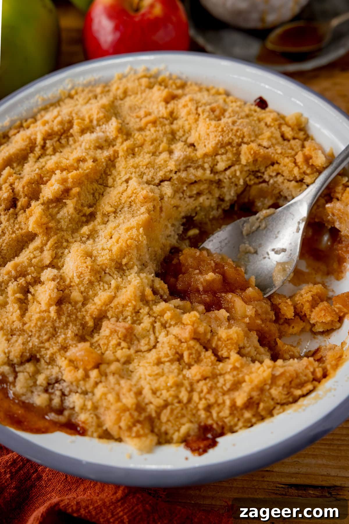 Irresistible Apple Crumble Perfection 4 A freshly baked apple crumble in a white ceramic dish, with a generous scoop already taken out. The dish sits invitingly on a wooden table, and a silver serving spoon is poised, scooping up some of the warm, cinnamon-spiced apple filling, highlighting the delicious textures of the dessert.