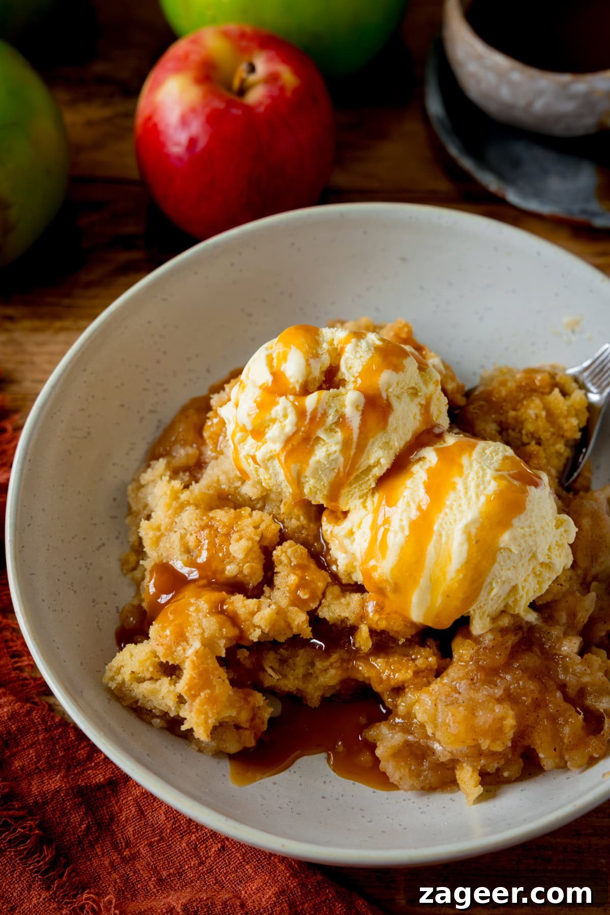 Irresistible Apple Crumble Perfection 5 A tall image showcasing a white bowl brimming with warm apple crumble, beautifully crowned with two scoops of melting vanilla ice cream. Golden caramel sauce is being skillfully drizzled over the ice cream. The exquisite dessert is presented on a wooden table, next to a dark orange napkin, with several fresh apples and a small bowl of caramel sauce artfully arranged nearby, creating a rustic and inviting scene.