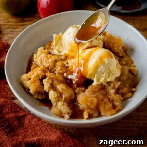A beautiful white bowl filled with golden apple crumble, adorned with two scoops of creamy vanilla ice cream. Caramel sauce is being drizzled enticingly over the ice cream, all set on a wooden table beside a dark orange napkin and fresh apples.