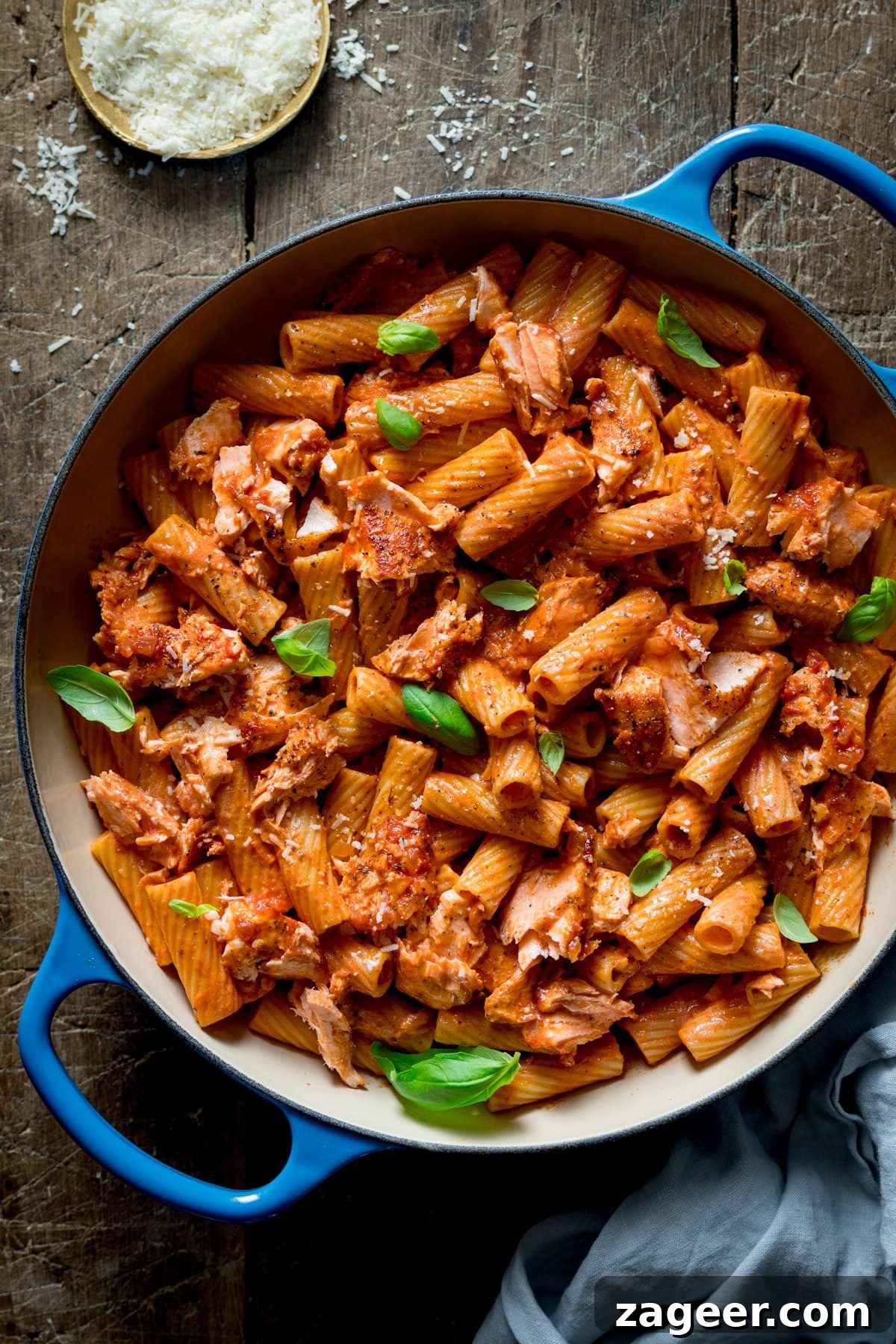 Blush Salmon Rigatoni 2 Overhead image of creamy tomato salmon and rigatoni in a blue cast iron pan on a wooden table. There is a dish of grated parmesan at the top of the image.