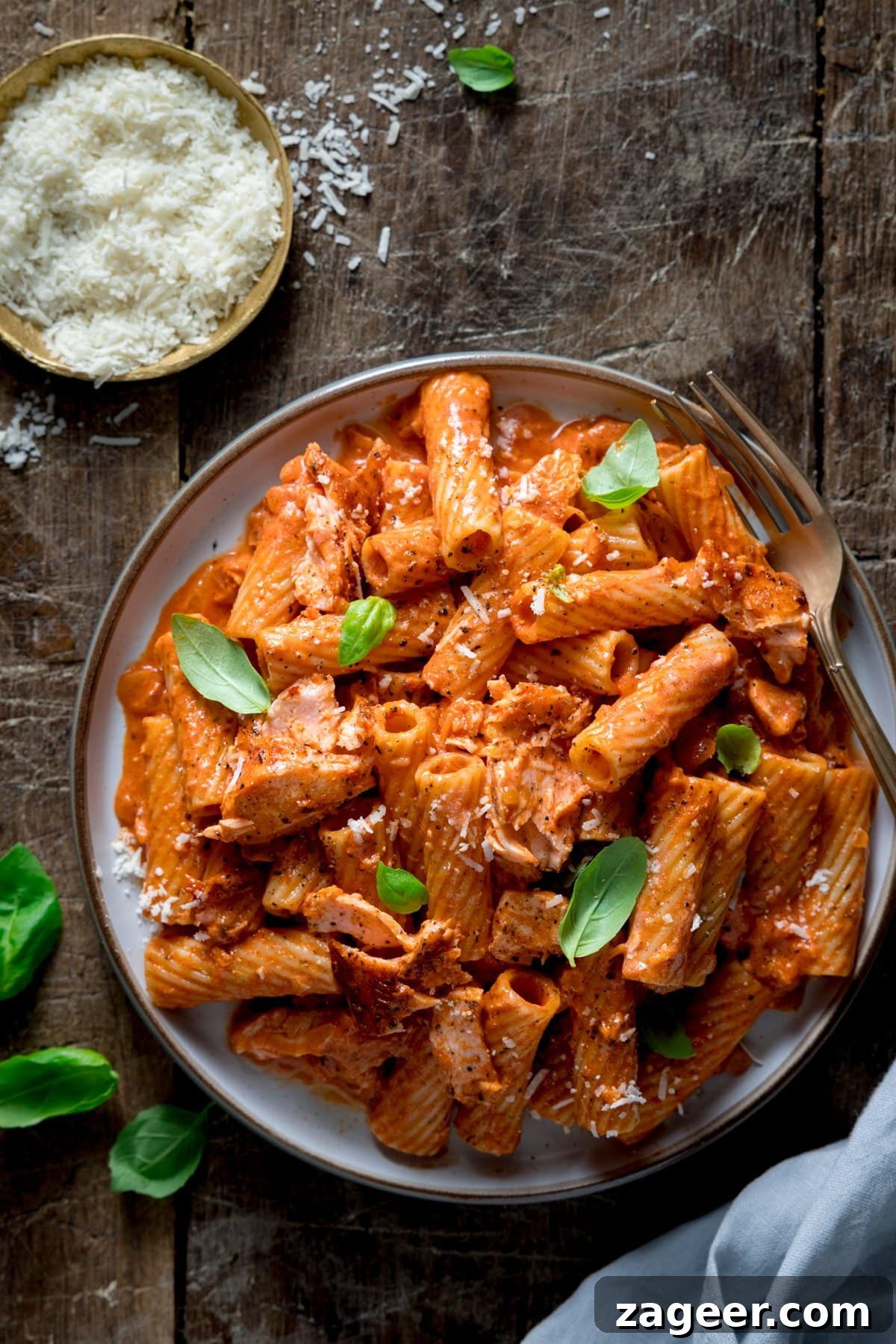 Blush Salmon Rigatoni 5 Overhead image of a plate of creamy tomato salmon pasta on a wooden table. There are basil leaves and a dish of grated parmesan around the plate.
