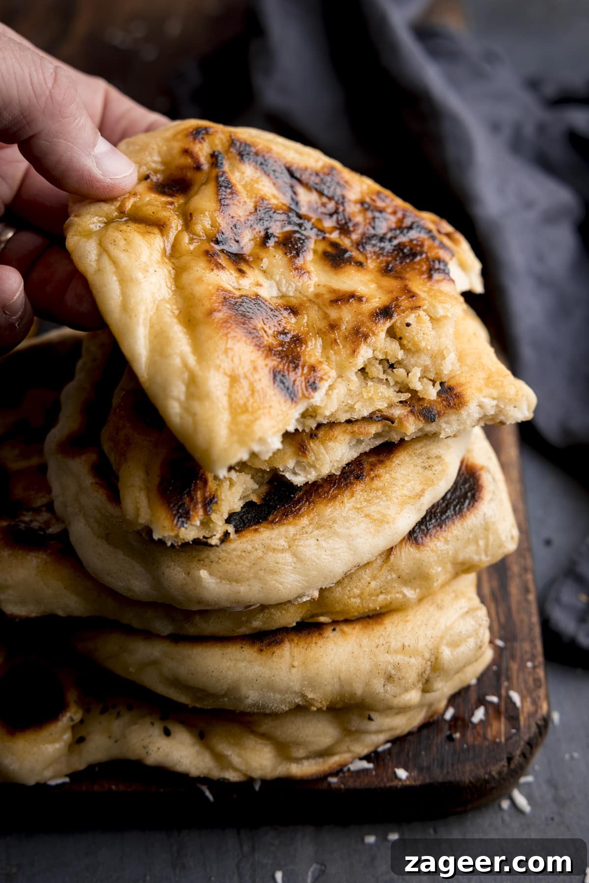 Homemade Peshwari Naan: Sweet & Fluffy Indian Bread 2 Tall image of a stack of peshwari naan breads on a wooden board against a blue background. There is a blue napkin in shot and a hand is taking the top naan.