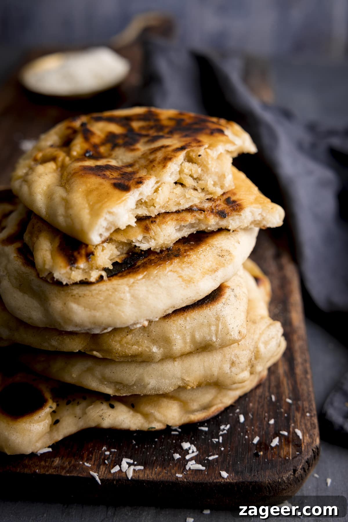 Homemade Peshwari Naan: Sweet & Fluffy Indian Bread 4 A stack of peshwari naan breads on a dark wooden board against a blue background. There is a blue napkin in shot. The top naan has been broken into two to show the filling.