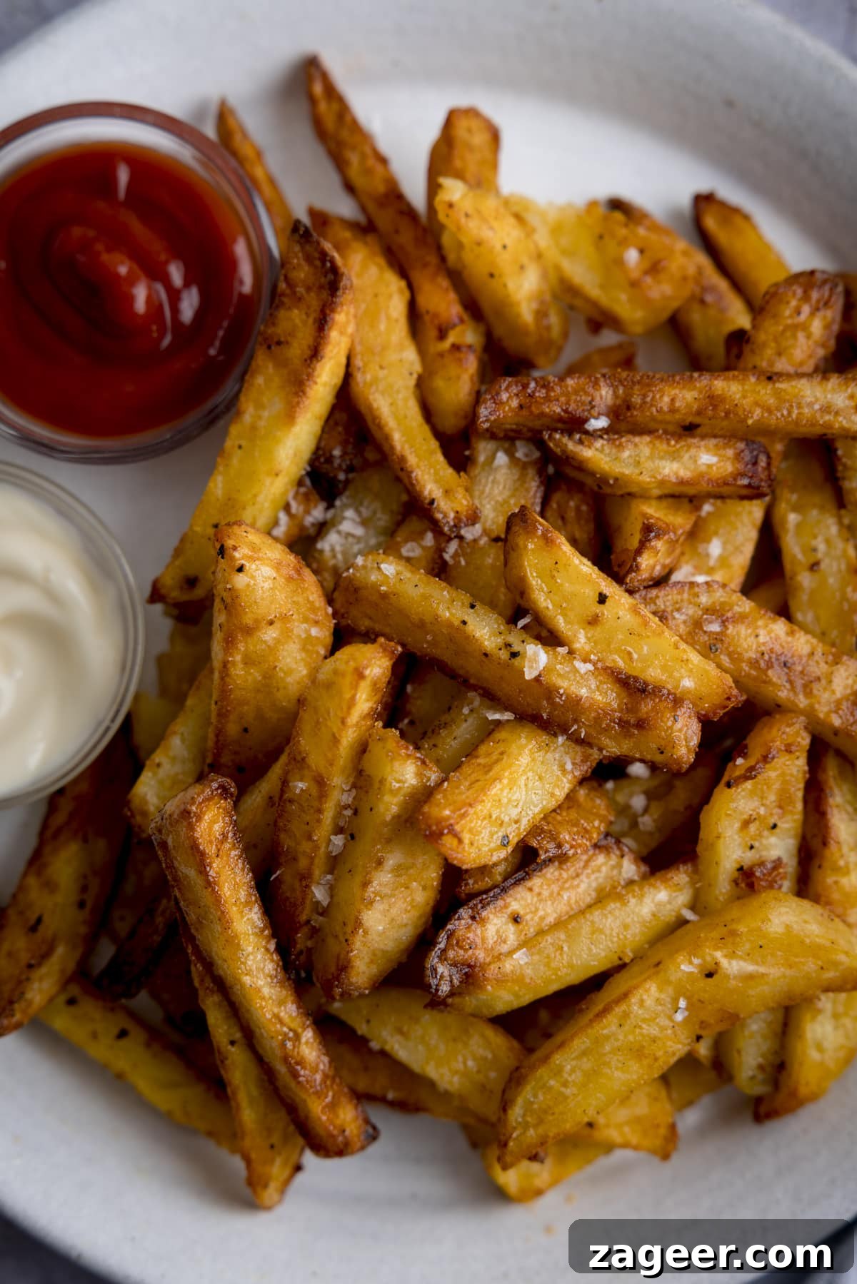 Perfectly Crispy Air Fryer Potato Chips 2 A tall overhead shot of perfectly golden, crispy air fryer chips on a white plate next to two small dipping bowls - one filled with vibrant red ketchup, one filled with creamy mayonnaise, ready for dipping.