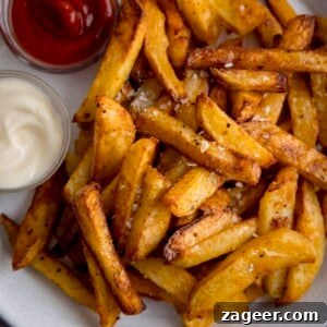 Perfectly Crispy Air Fryer Potato Chips 7 A square overhead shot of Air Fryer Chips, perfectly sprinkled with salt and black pepper, served on a crisp white plate alongside small bowls of ketchup and mayonnaise for dipping.
