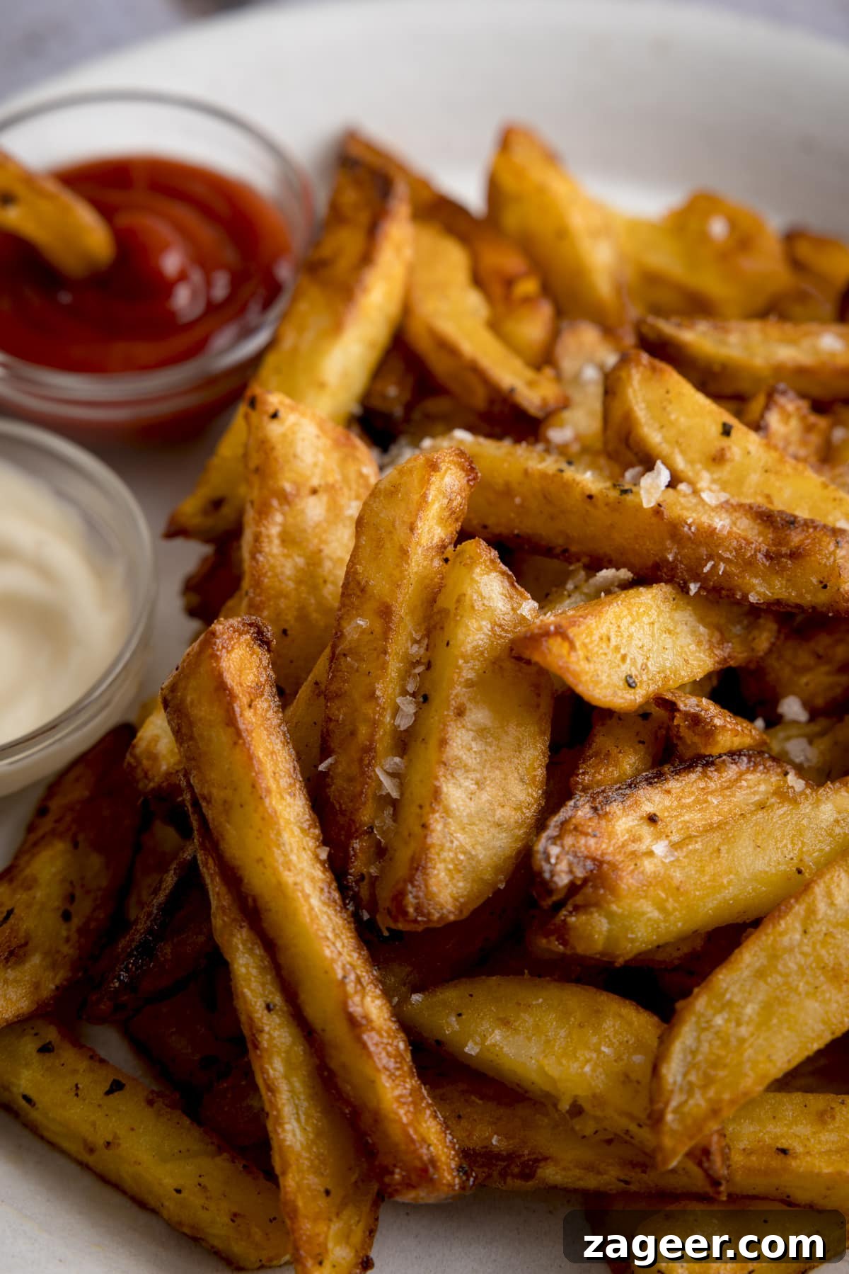 Perfectly Crispy Air Fryer Potato Chips 5 Close up of a mound of golden Air Fryer Chips sprinkled with salt and black pepper, on a white plate with a small bowl of ketchup with a chip dunked in on the side and a small bowl of mayonnaise, showcasing the perfect crispness.