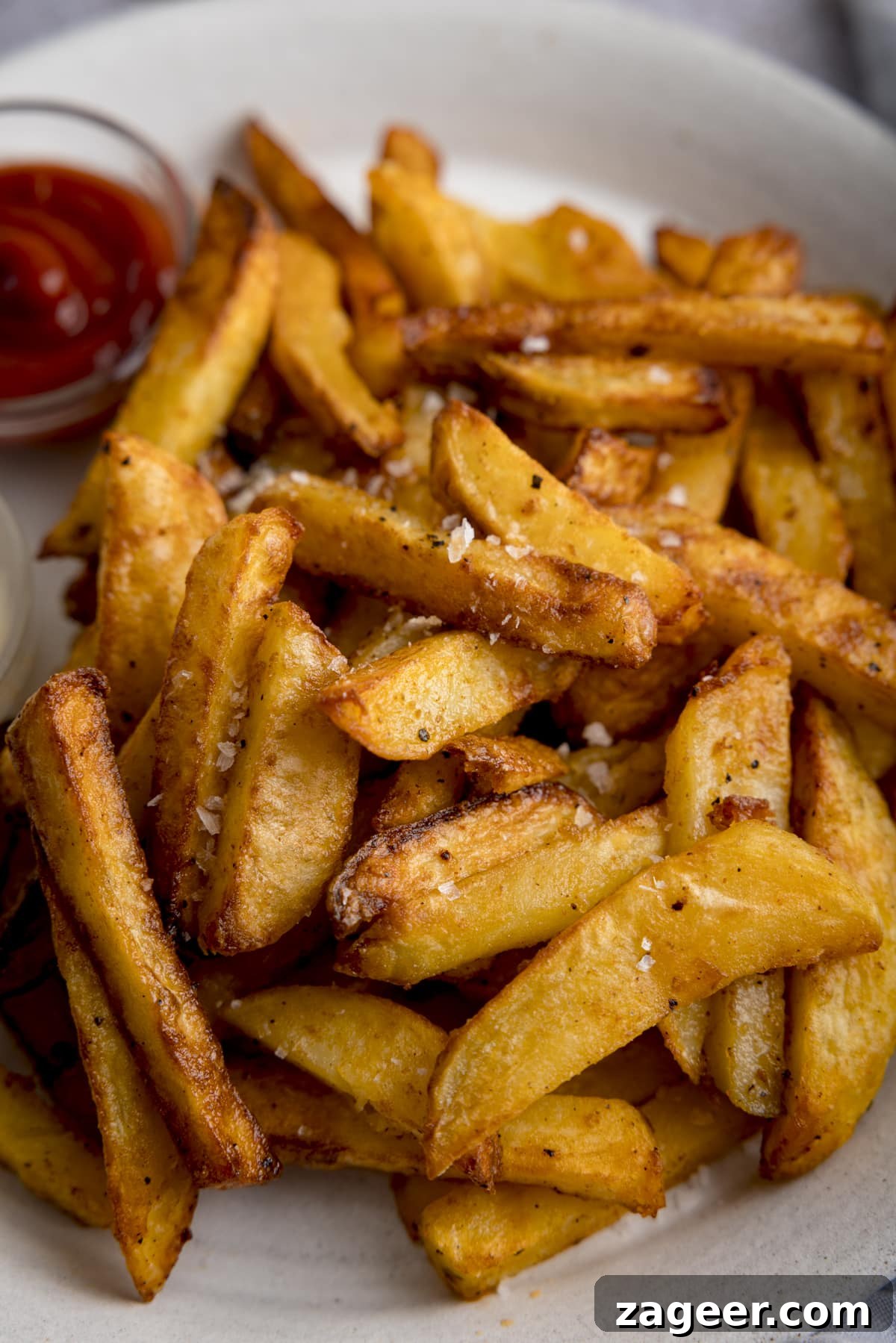 Perfectly Crispy Air Fryer Potato Chips 6 A tall close up shot of a mound of golden Air Fryer Chips sprinkled with salt and black pepper, on a white plate with a small bowl of ketchup on the side and a small bowl of mayonnaise, highlighting their crisp texture.