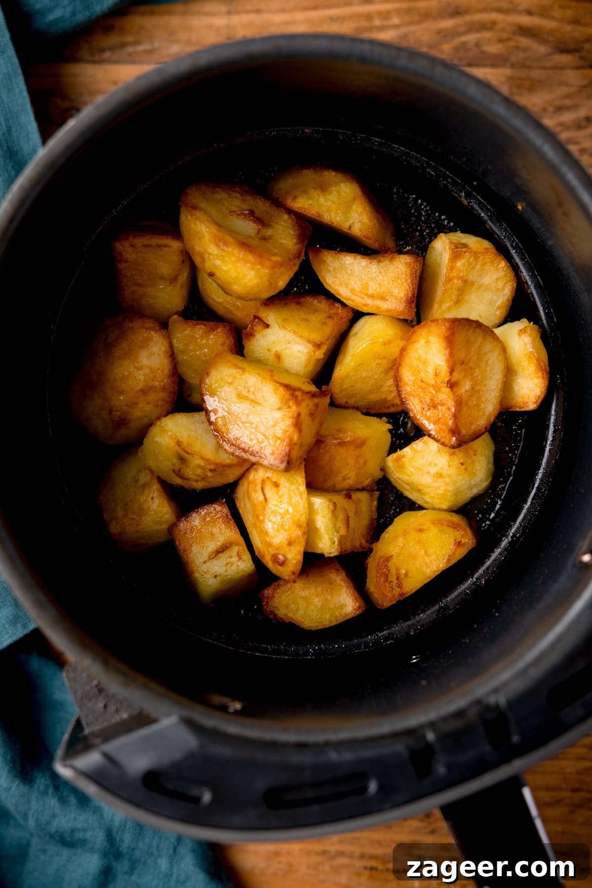 Ultra-Crisp Air Fryer Roast Potatoes 5 Tall overhead image of perfectly cooked air fryer roast potatoes piled up inside an air fryer basket. The air fryer basket is shown on a wooden table next to a folded green napkin, emphasizing the readiness of the delicious side dish.