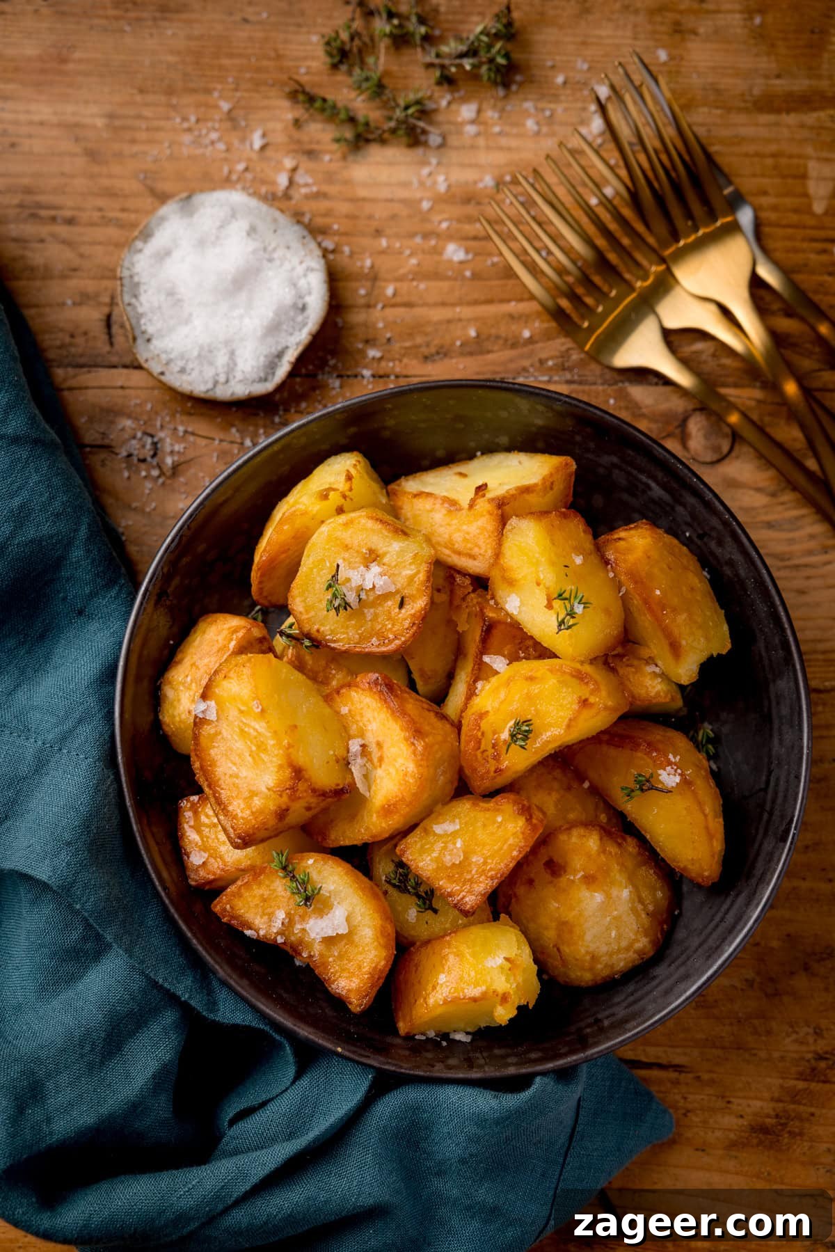 Ultra-Crisp Air Fryer Roast Potatoes 6 Tall overhead image of golden and crispy air fryer roast potatoes piled up in a dark serving bowl. The potatoes are garnished with fresh thyme sprigs and sprinkled with flaky sea salt. The bowl is set on a rustic wooden table next to a green napkin, a small dish of salt, and some gold-colored forks, creating an inviting tableau.