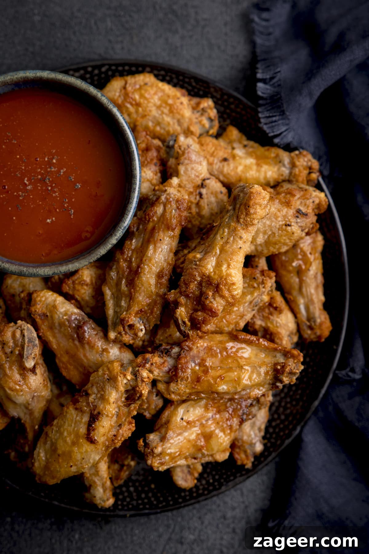 Perfectly Crispy Air Fryer Wings 2 A tall shot of Air Fryer Crispy Chicken Wings piled up on a black speckled plate, with a large black bowl of buffalo sauce and seasoning. The plate is on a mottled dark grey background with a dark blue napkin.