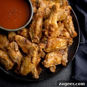 Perfectly Crispy Air Fryer Wings 6 A square overhead shot of Air Fryer Crispy Chicken Wings piled up on a black speckled plate, with a large black bowl of buffalo sauce and seasoning. The plate is on a mottled dark grey background with some dark blue napkins to the side.