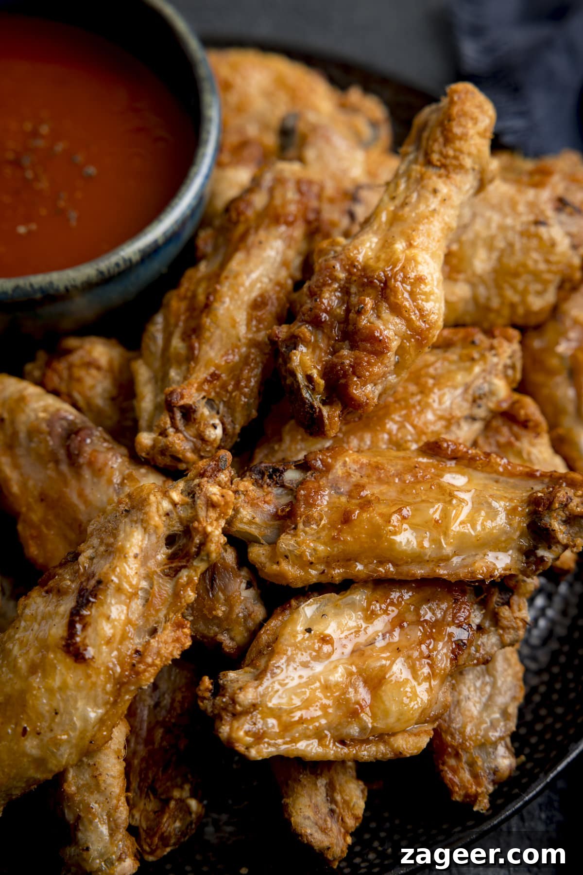 Perfectly Crispy Air Fryer Wings 4 A close up shot of Air Fryer Crispy Chicken Wings piled up on a black speckled plate, with a large black bowl of buffalo sauce, sprinkled with seasoning. The plate is on a mottled dark grey background with a dark blue napkin in the background.