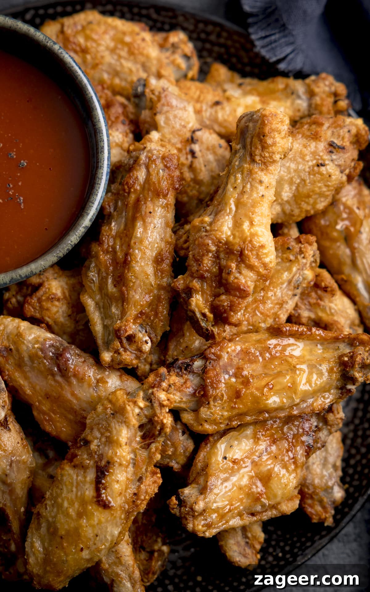 Perfectly Crispy Air Fryer Wings 5 A tall close up shot of Air Fryer Crispy Chicken Wings piled up on a black speckled plate, with a large black bowl of buffalo sauce and seasoning. The plate is on a mottled dark grey background with a dark blue napkin to the side.