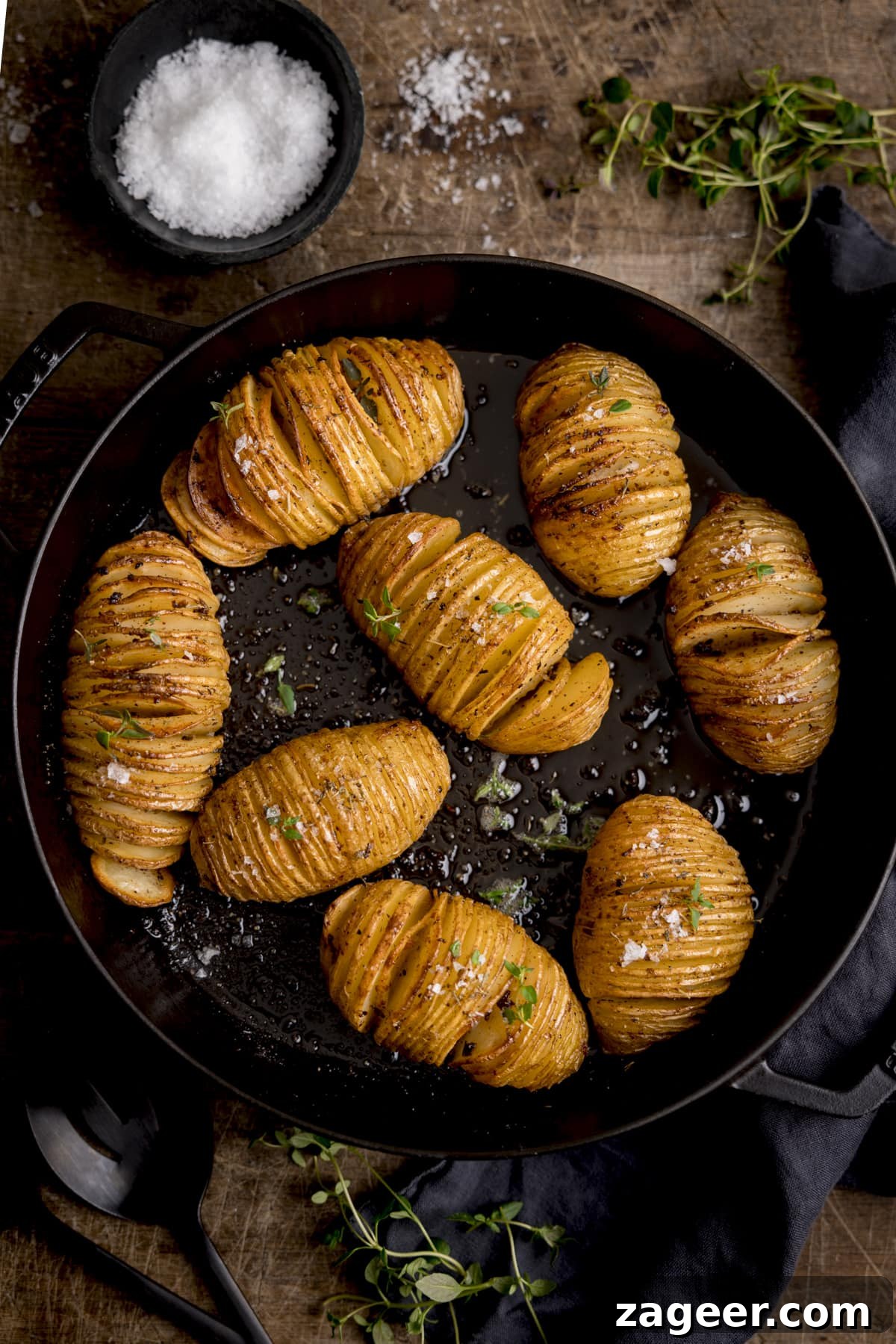 Crispy Accordion Potatoes 2 Overhead image of hasselback potatoes in a dark cast iron pan, topped with fresh thyme leaves and Maldon salt. The pan is on a wooden table and there is a small bowl of Maldon salt and some thyme leaves around the pan.