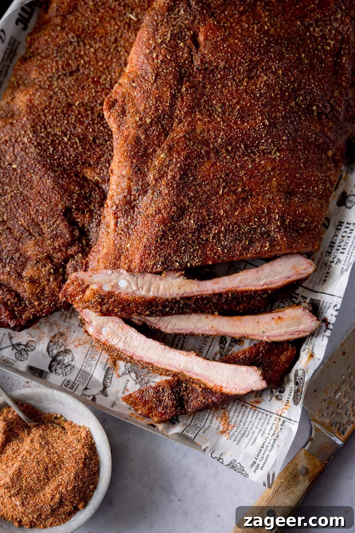 Smoky Dry Rub BBQ Ribs 2 A tall, close-up image of dry rub BBQ ribs. Two racks of beautifully smoked ribs are laid out on a silver tray lined with newspaper. The central rack is cut, with three ribs separated to showcase their juicy interior and perfect smoke ring. In the bottom left, a light grey bowl holds the vibrant dry rub seasoning with a silver teaspoon. A large silver knife with a wooden handle rests angled on the bottom right. All elements are set against a light grey background.
