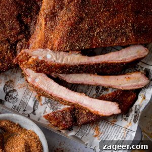A square, close-up image of dry rub BBQ ribs. Two racks of ribs are laid out on a silver tray lined with newspaper. The central rack is cut, with three ribs separated. A bowl of dry rub and a silver knife are visible in the foreground.