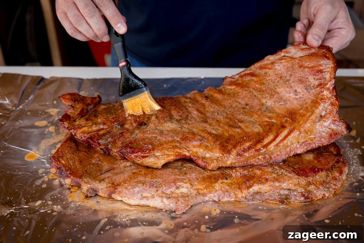 Smoky Dry Rub BBQ Ribs 7 A prep shot of BBQ Dry Rub Ribs. The ribs are laid out on a sheet of silver tinfoil on a white surface. Two hands are visible; one holds an end of the ribs, while the other holds a brush, applying the mop sauce evenly over the surface.
