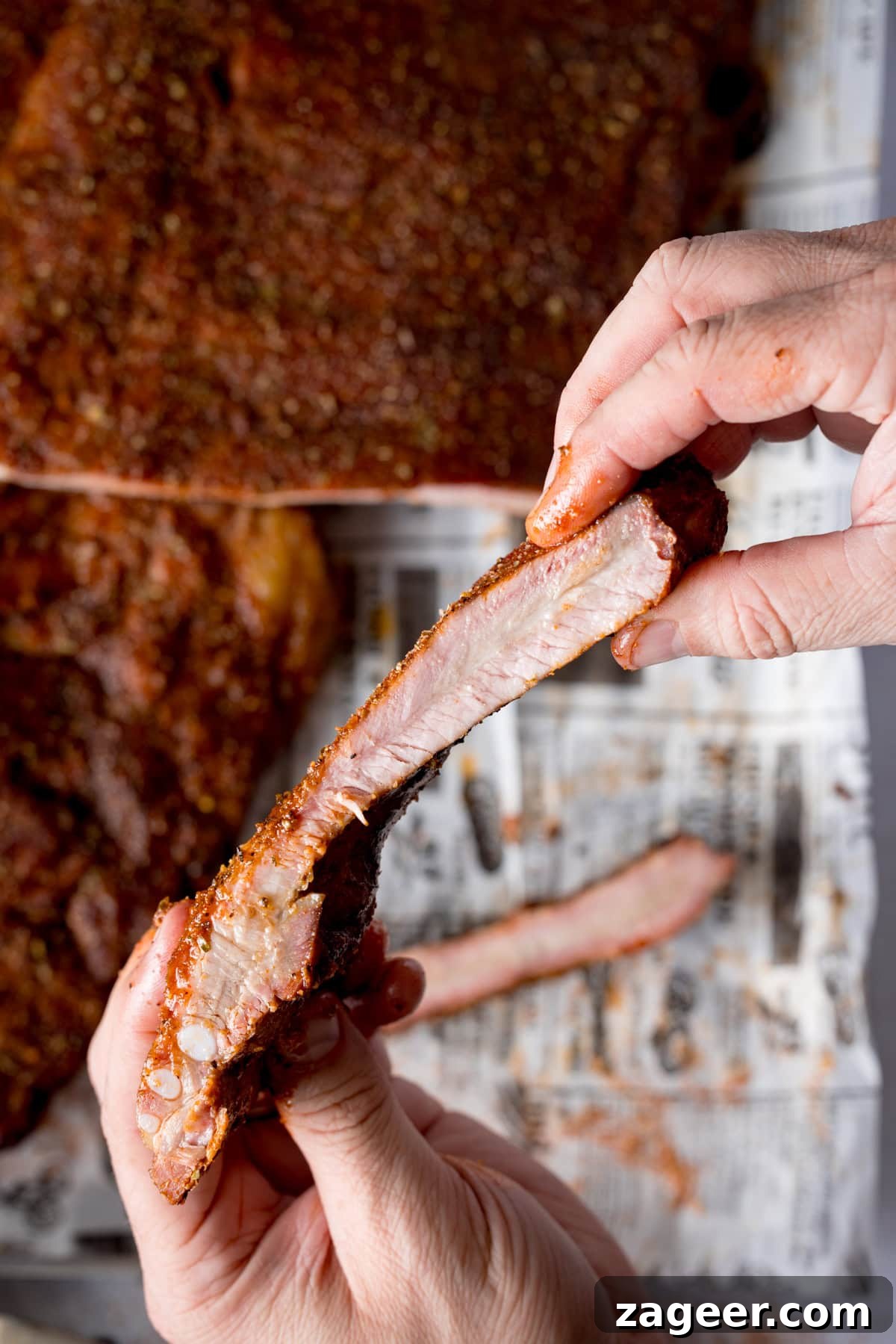 Smoky Dry Rub BBQ Ribs 9 A tall, close-up image of dry rub BBQ ribs. Two racks of beautifully cooked ribs are arranged on a silver tray lined with newspaper. The central rack is cut, with two ribs separated to reveal their tender interior and perfect smoke ring. In the foreground, two hands gently pick up a single rib, showcasing its succulence and the juicy meat pulling away from the bone. All set against a light grey background.