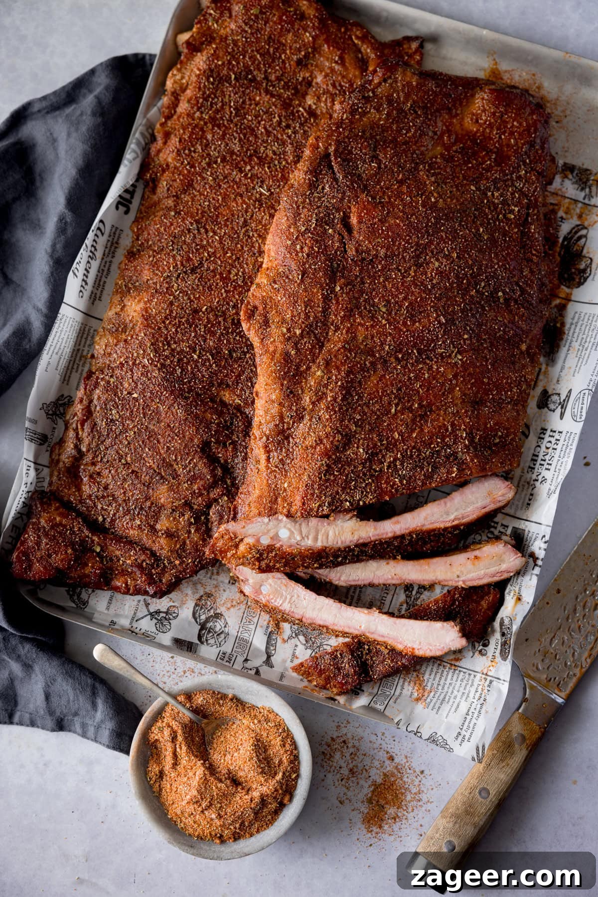 Smoky Dry Rub BBQ Ribs 10 A tall, overhead image of dry rub BBQ ribs. Two racks of the ribs are beautifully laid out on a silver tray, lined with newspaper. The central rack is cut, with three ribs separated to showcase their tenderness and smoky appearance. In the bottom left, a slate grey napkin is tucked underneath the tray, while the dry rub seasoning sits in a light grey bowl with a silver teaspoon in the bottom center. A large silver knife with a wooden handle is angled on the bottom right. This appealing arrangement is set on a light grey background.