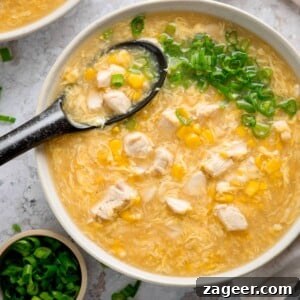 Overhead square shot of a white bowl filled with chicken and sweetcorn soup. The soup is topped with spring onions and there is a black spoon sticking out. There are some spring onions also in shot.