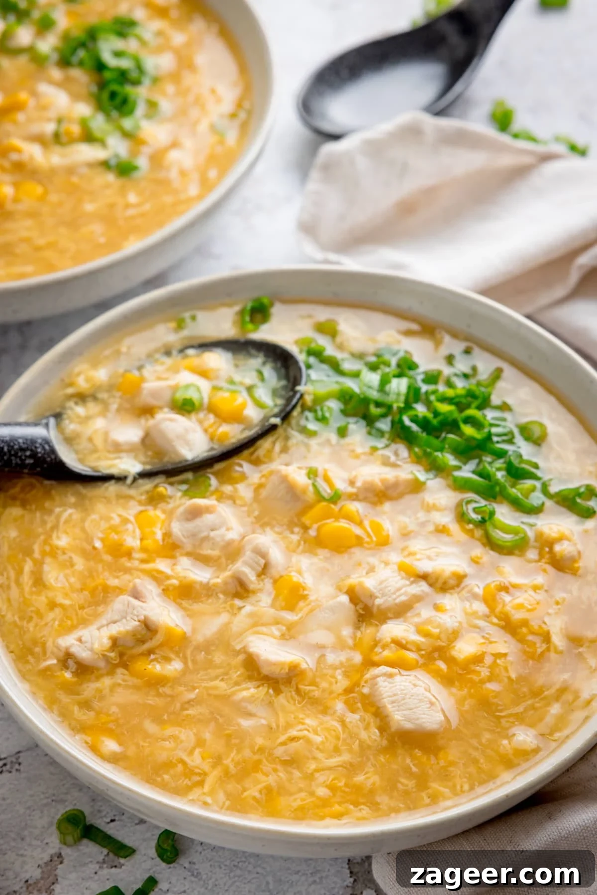 Savory Chicken and Sweet Corn Delight 4 Bowl of chicken and sweetcorn soup with spring onions on top and a black spoon sticking out. There is a further bowl of soup in the background, along with a another black spoon and a cream napkin.