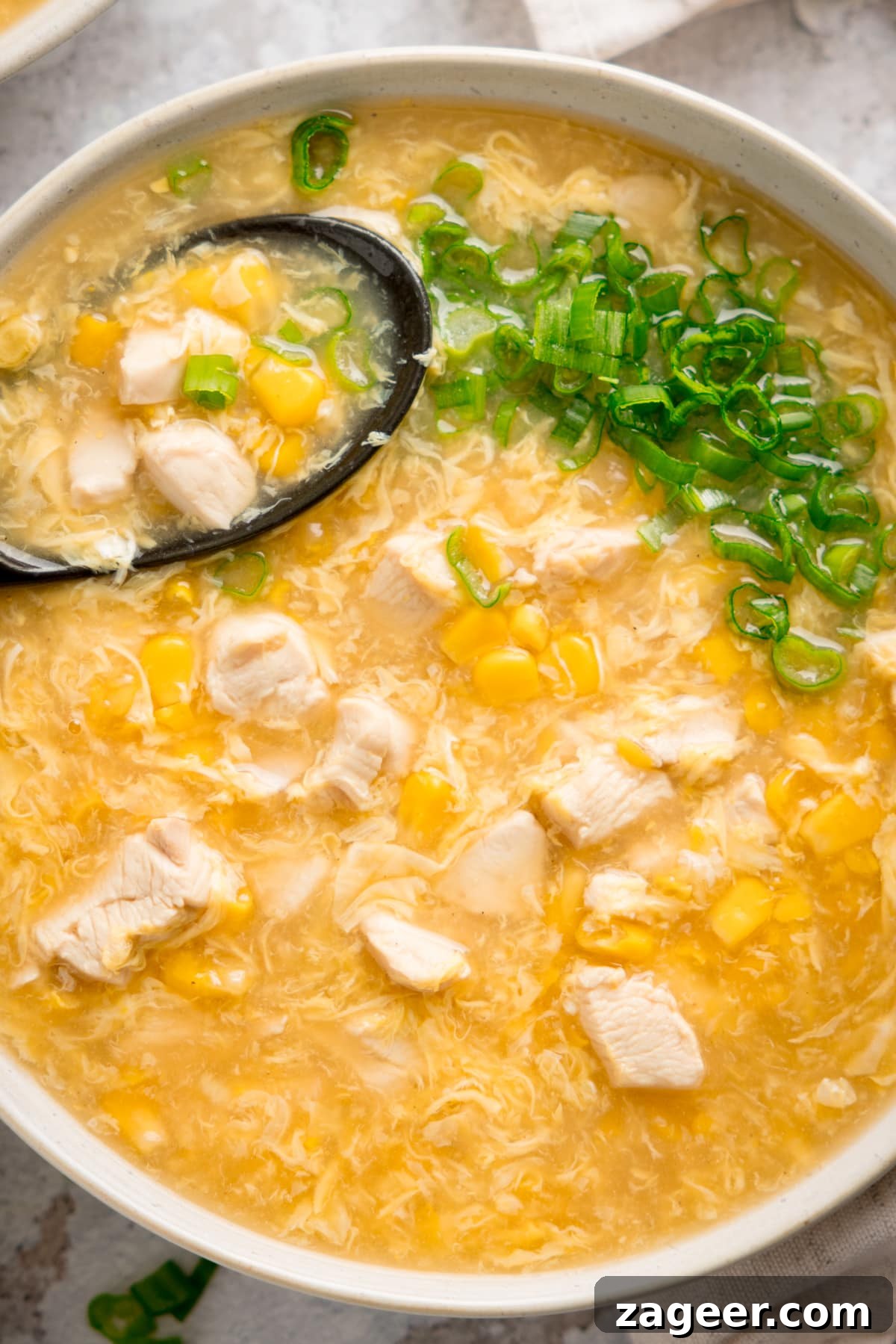Savory Chicken and Sweet Corn Delight 5 Close up overhead shot of a white bowl filled with chicken and sweetcorn soup. There are chopped spring onions on top and a black spoon sticking out of the soup.