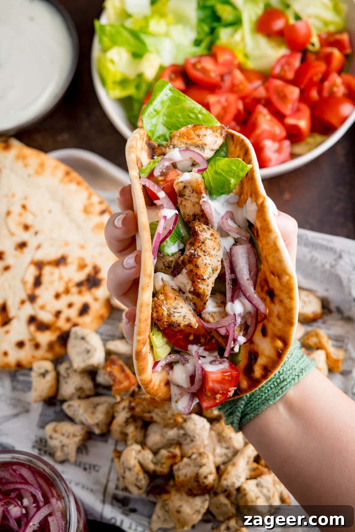 Lemon Herb Air Fryer Greek Chicken 6 Overhead shot of an Air Fryer Greek Chicken wrap being held by a hand, showcasing the delicious chicken, salad, and feta dressing stuffed into a flatbread, with other cooked ingredients on a tray below.
