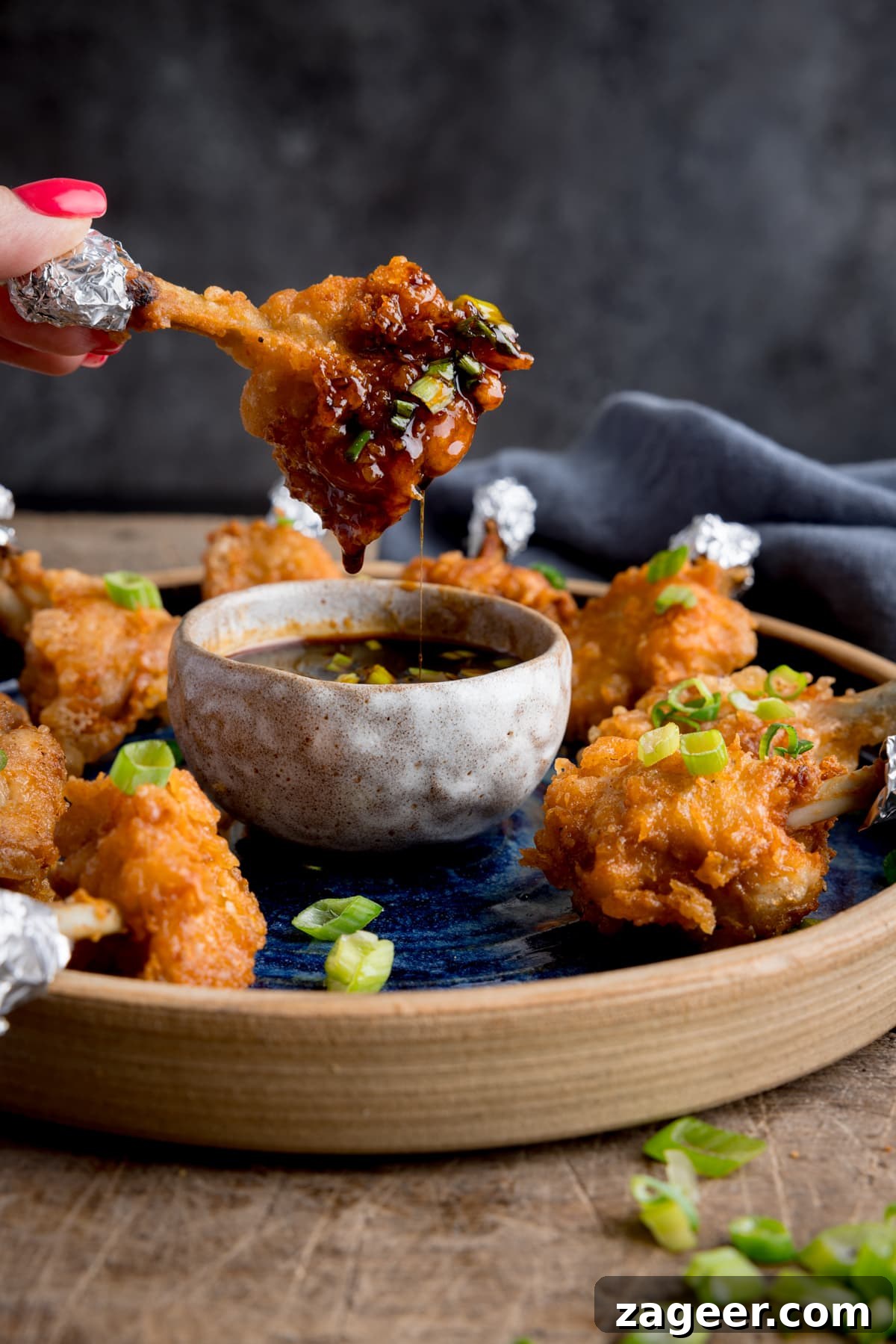 Golden Crispy Chicken Lollipops 2 A plate of chicken lollipops with a bowl of spicy dipping sauce. One of the wings is being dipped into the sauce and is dripping. The wing is being held by fingers with pink-painted fingernails.