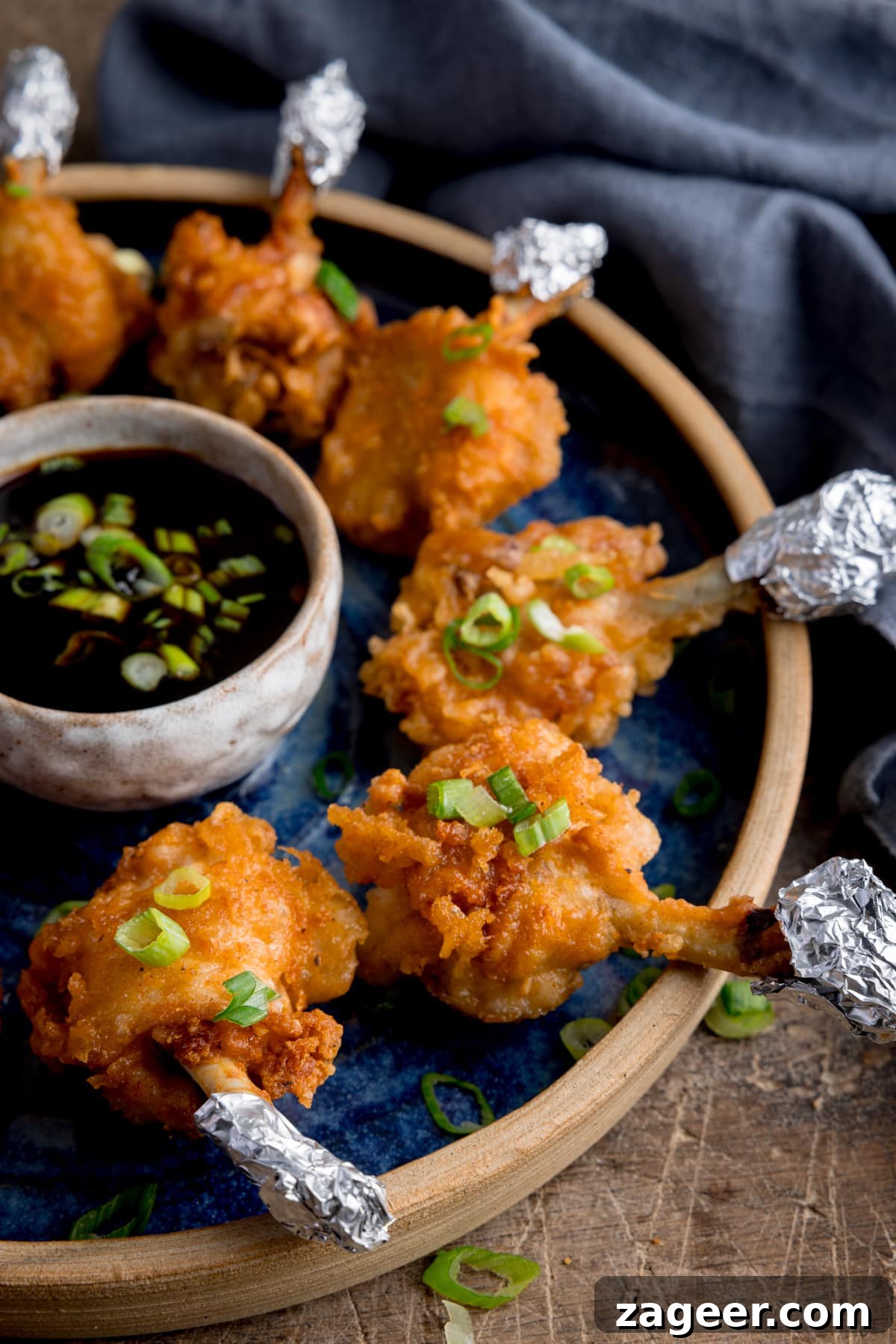 Golden Crispy Chicken Lollipops 7 Close up side image of chicken lollipops arranged in a circle on a blue plate with a dipping bowl of sticky spicy sauce int he middle. The lollipops and sauce have been sprinkled with chopped spring onions and the plate is in a wooden table next to a blue napkin.