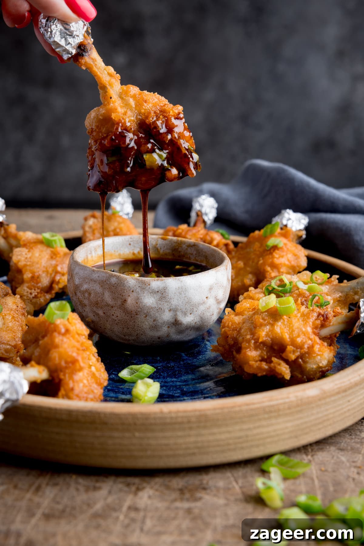 Golden Crispy Chicken Lollipops 6 A plate of chicken lollipops with a bowl of spicy dipping sauce. on a wooden table. One of the wings is being dipped into the sauce and is dripping. The wing is being held by fingers with pink-painted fingernails.