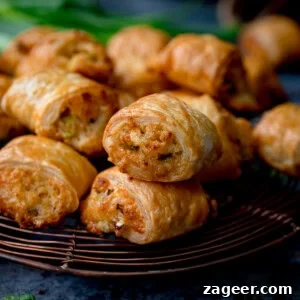 A close-up of golden-brown vegetarian sausage rolls stacked on a wire cooling rack, with a blurred background of more rolls and green herbs, highlighting their delicious appearance.