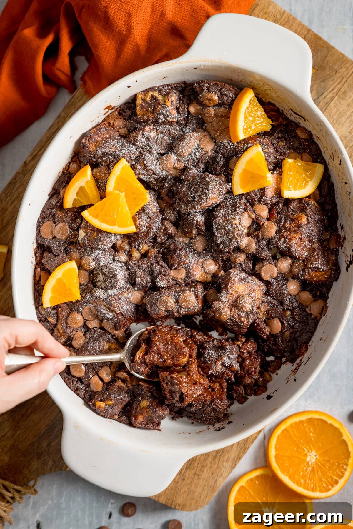 Overhead image of chocolate orange bread and butter pudding in a white dish, set on a wooden board. The pudding has orange slices on top and a spoonful is being taken. There are orange slices and an orange napkin around the dish.