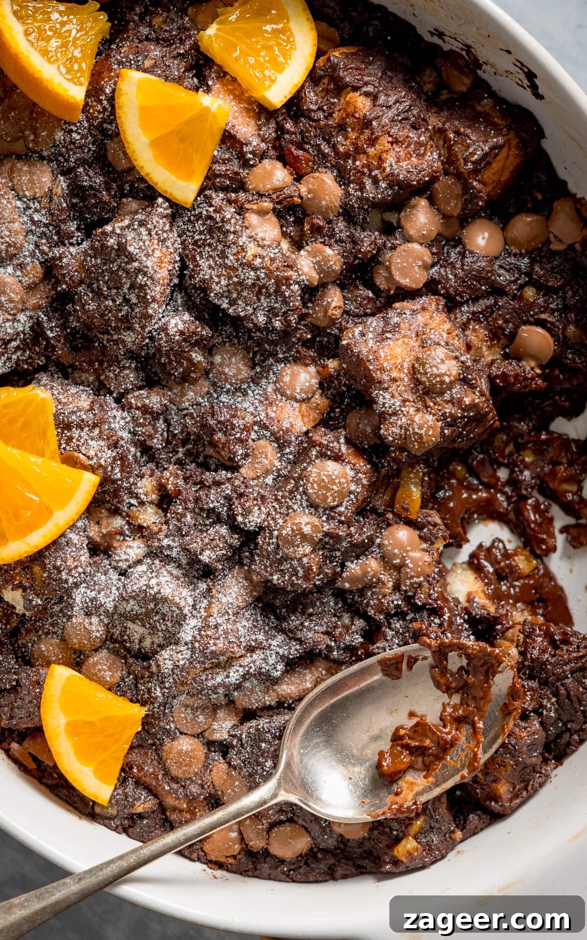 Close up overhead image of chocolate orange bread and butter pudding in a white dish. The pudding has orange slices on top and a spoonful is being taken.