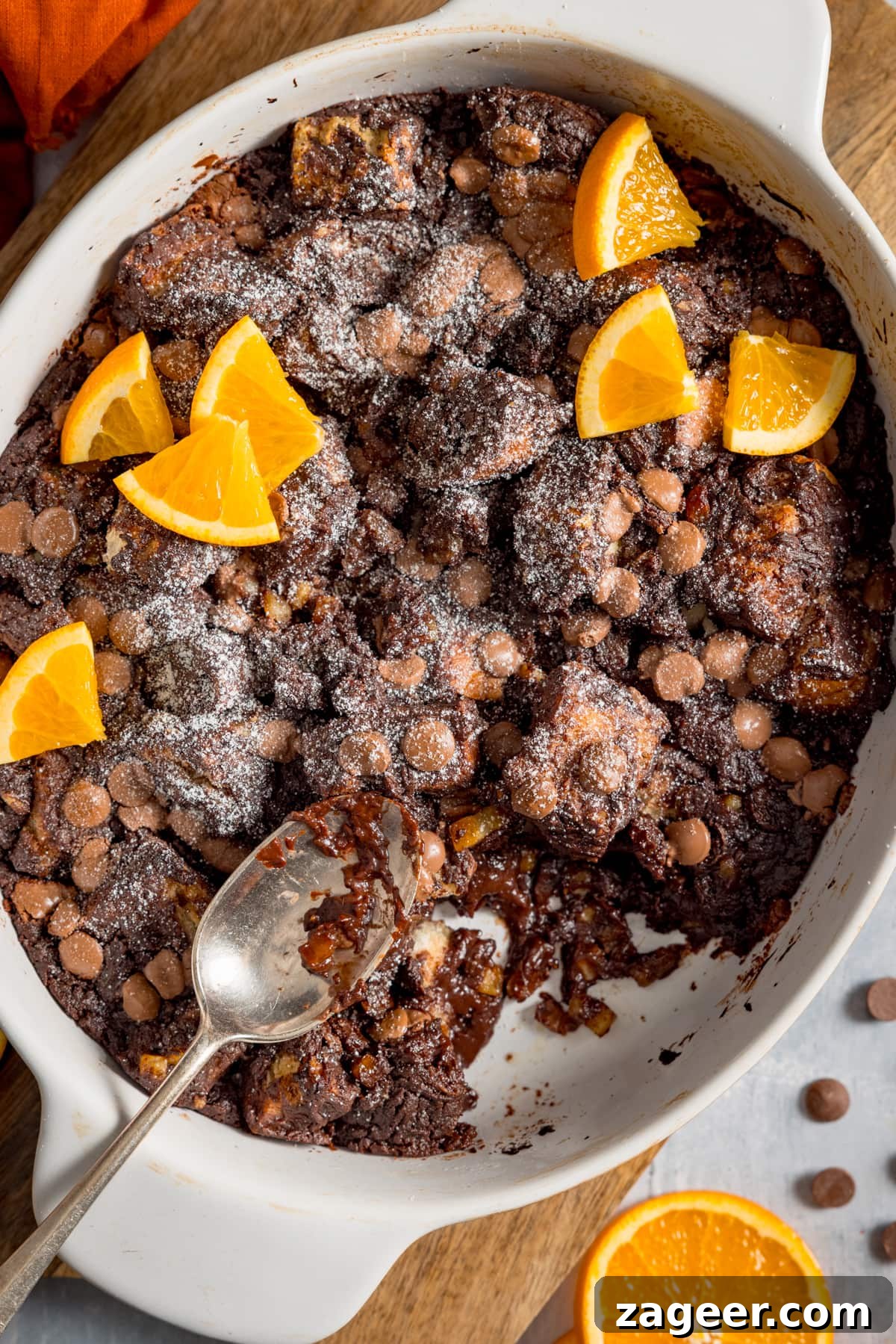 Tall image of chocolate orange bread and butter pudding in a white dish, set on a wooden board. The pudding has orange slices on top and a spoonful is being taken.