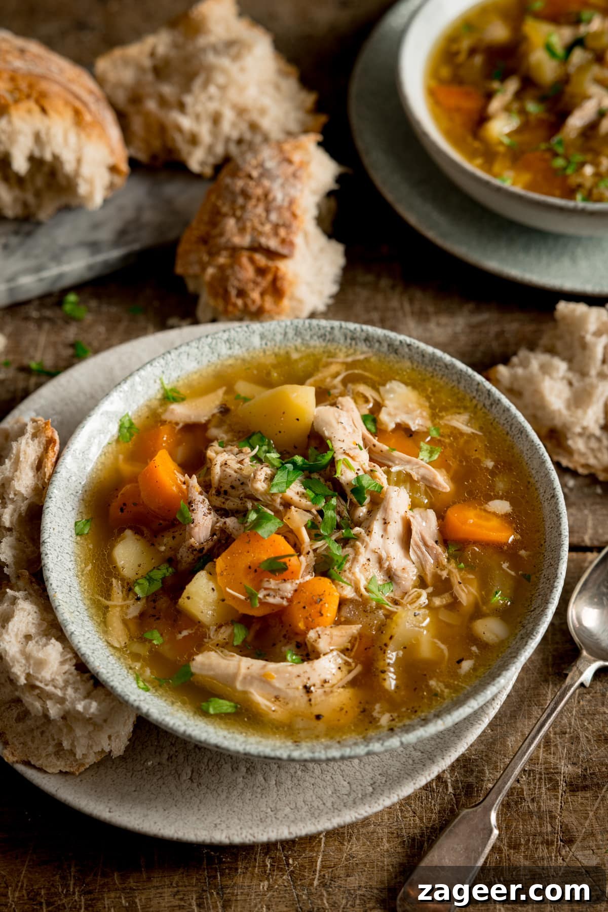 Hearty Slow Cooker Chicken and Veggie Bowl 2 A tall, inviting image of slow cooker chicken and vegetable soup served in a light bowl, resting on a rustic wooden table. A chunk of artisan bread is nestled beside the bowl, alongside a spoon, emphasizing the comforting nature of the meal. Another bowl of soup and a torn loaf of bread are visible in the background, setting a warm, homely scene.