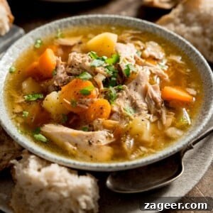 Square image of chicken and vegetable soup in a light bowl. The bowl is sat on a plate. There is a chunk of bread and a spoon nestled between the bowl and plate.