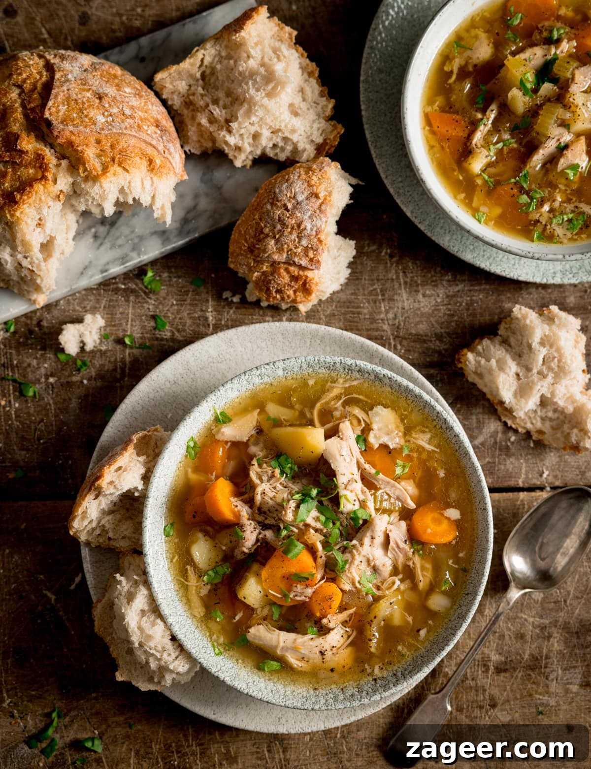 Hearty Slow Cooker Chicken and Veggie Bowl 4 An overhead image capturing the warmth of slow cooker chicken and vegetable soup in a light bowl, presented on a wooden table. A slice of crusty bread and a spoon enhance the rustic appeal. Another bowl and fresh bread are subtly visible in the soft-focus background.